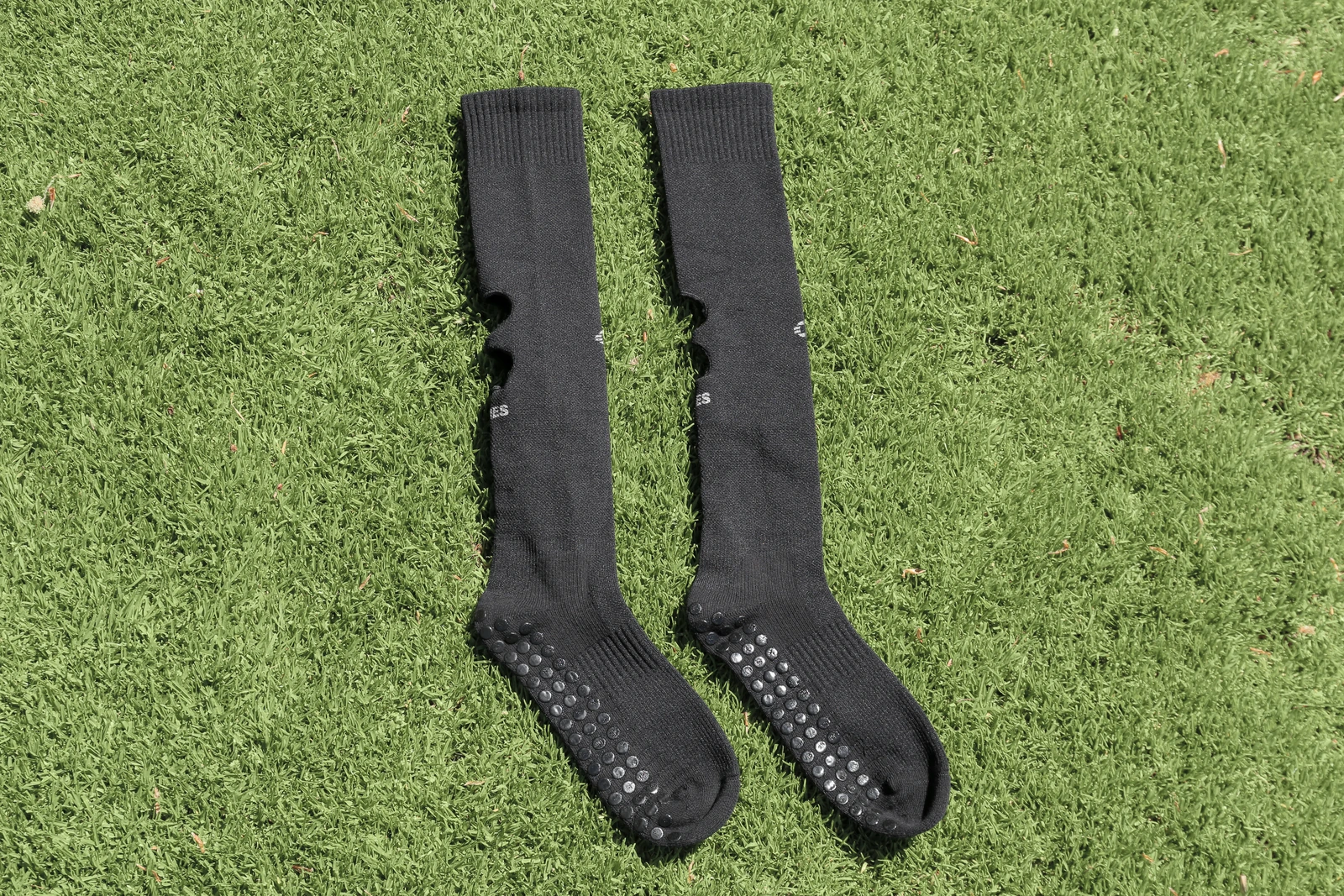 Zwei schwarze HOLIES Grip Cut Socks auf Rasen, mit sichtbarem Bündchen, Schaft und Fußteil samt Grip-Punkten.